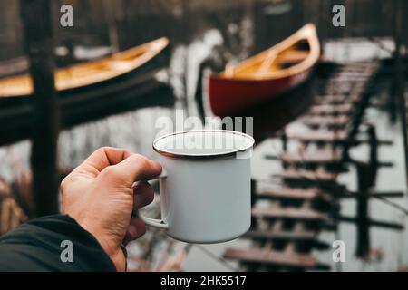 A tin hiking cup in hand with canoe boats in the background, point of view shot. Paddling lifestyle in springtime or autumn season Stock Photo