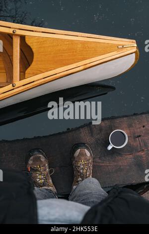 Front part of a canoe, tin cup of coffee and feet in trekking boots, point of view shot. Directly above view of somebody standing next to a beautiful Stock Photo