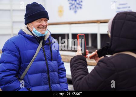 Senior Caucasian woman photographing her gray-haired smiling wearing protective face mask senior man husband at smart phone. Seniors using new technol Stock Photo