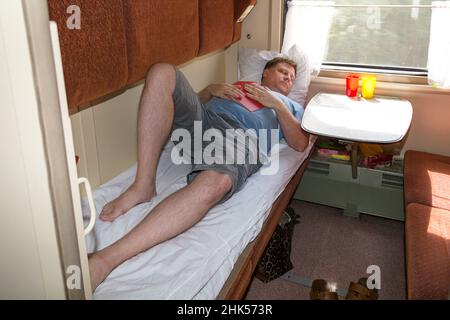 An adult blond man of 40-45 years old is lying down train compartment reading a book. Stock Photo