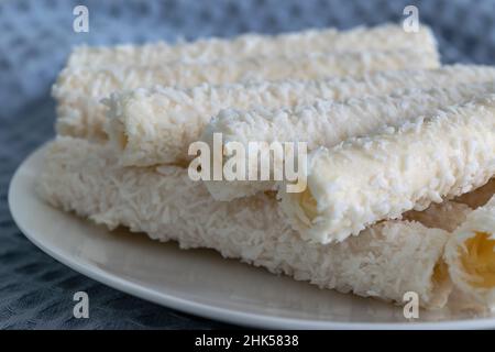 Sweet wafer rolls in coconut flakes on a white porcelain plate and a gray cotton kitchen towel. Horizontal photo. Stock Photo