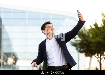Asian tourist businessman arrived at a business conference takes photos near the airport and communicates by video with colleagues using a smartphone Stock Photo