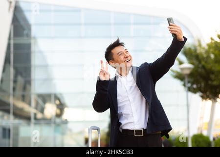Asian tourist businessman arrived at a business conference takes photos near the airport and communicates by video with colleagues using a smartphone Stock Photo