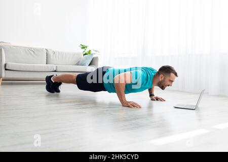 Cheerful millennial muscular european man doing push ups and watching online lesson on computer in living room Stock Photo