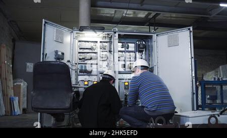 Installers of electrical equipment at factory. Clip. Two installers in helmets look at problems in electrical equipment. Installation of industrial eq Stock Photo