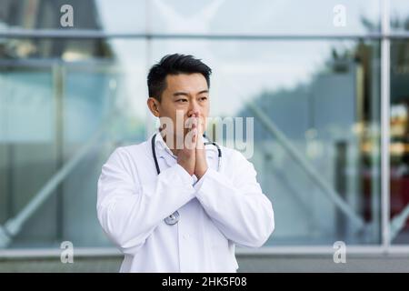 portrait Asian doctor tired after work, puts his hands to his face hoping for a good result on the street near the clinic Stock Photo