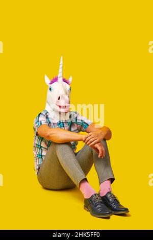 a young man, wearing a unicorn mask, and colorful clothes, is sitting on the floor on a yellow background with some blank space on top Stock Photo