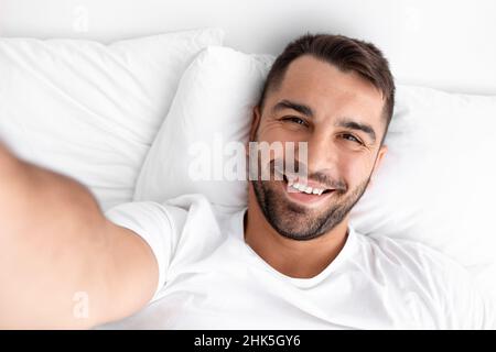 Smiling glad millennial muscular caucasian man waking up taking selfie on white bed on soft pillow in bedroom Stock Photo