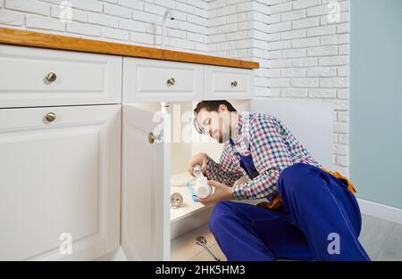 Young serious professional plumber installs new siphon under sink in kitchen of apartment. Stock Photo