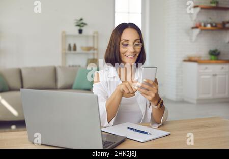 Happy woman using mobile phone while sitting at desk with laptop and notebook at home Stock Photo