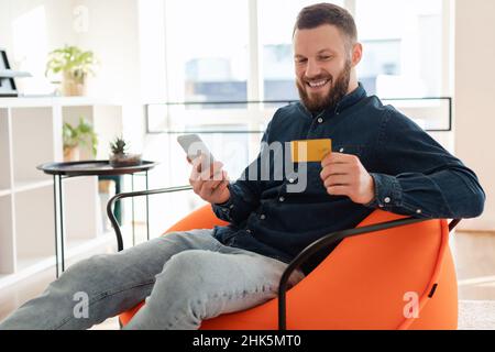 Cheerful Man Shopping Using Phone And Credit Card Sitting Indoors Stock Photo