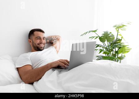 Satisfied young muscular caucasian man typing on computer lies on bed in bedroom interior Stock Photo