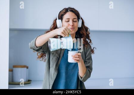 Curly-haired young woman with loose hairstyle dances to loud music in wireless headphones with coffee in cup and coffeepot Stock Photo