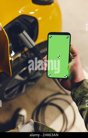 Man charging electric vehicle with cable looking at app on mobile phone, green screen Stock Photo
