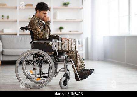 Upset young male looking at camera with sad look. He is sitting in invalid chairing wearing military uniform. Isolated on grey background. Copy space Stock Photo