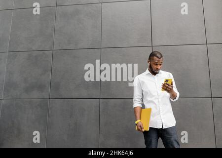 Portrait of african american male freelancer using smartphone, surprised with received message. Young adult stylish businessman standing near the Stock Photo