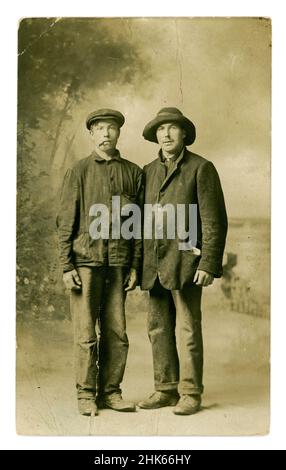 Original, clear, Edwardian era postcard of 2 poor working class men, friends, characters, possibly itinerant labourers, also known as navvies. The men are wearing dirty workwear, jackets, hat and flat cap, one smoking a cigar (he looks like he could be trouble, a rogue!), the other is very shabby looking wearing a jacket with the buttons undone and a crumpled hat who seems to have his lunch in his pocket. They are photographed in their scruffy working clothes as if they have been hurriedly photographed after working outside.  U.K. circa 1905. Stock Photo