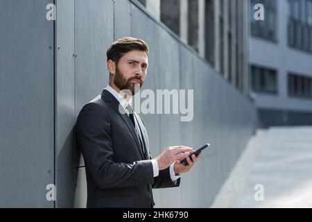 Caucasian bearded employee on urban background office building texting Messages, read good news mobile phone. Business man standing a formal suit mess Stock Photo