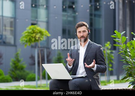 a man quarrels and frustrated talking on video, using a laptop and a headset with a microphone, an angry supporter sitting in a public place near the Stock Photo