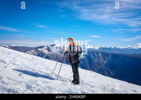 Girl on hike goes up the top of the snowy mountain. In the background the snow-covered mountain ranges. On a beautiful winter day. Stock Photo
