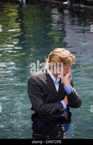 Young man wearing business suit, standing waist deep in water, hand on his face Stock Photo