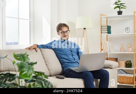Young attractive man sitting on sofa at home working online on laptop or using internet. Stock Photo