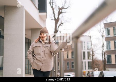 Portrait of confident overweight woman in warm hat and jacket standing near railing of building at urban street in cloudy autumn day, Stock Photo