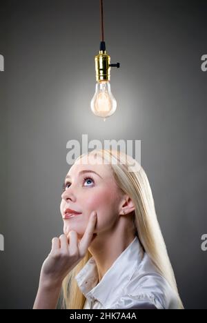 Young blonde woman, hand on chin, contemplates lit Edison light bulb Stock Photo