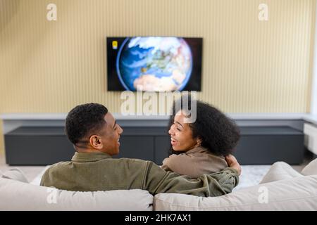 Back view of the caucasian couple in love sitting at the sofa and watching TV with pleasure smiles. Multiracial man embracing his lovely wife and looking at each other. Home concept Stock Photo
