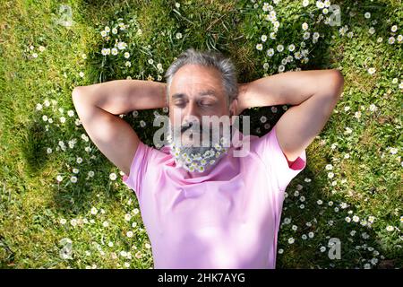 older man with a beard decorated with flowers lying on the grass on a sunny day Stock Photo
