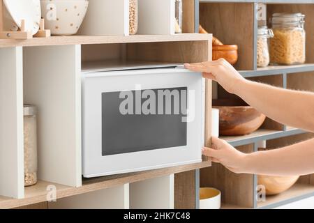 Young woman opening microwave oven on shelf unit in kitchen, closeup Stock Photo