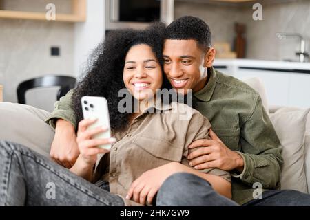 Multiracial couple with smartphone on the couch spending leisure time in social networks, scrolling feed, watching videos. Black guy embracing with his girlfriend Stock Photo