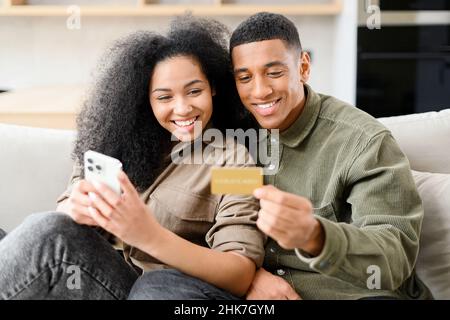 Online transaction, payment. Multiracial man holding credit card and using smartphone for shopping with his lovely woman. Dark skinned couple making purchases online Stock Photo