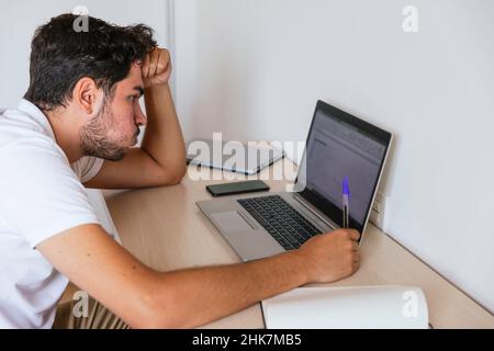 Side view of a young man puffing through his mouth, worried, sitting at a desk with his hand on his head with a computer taking notes. Concept: telewo Stock Photo
