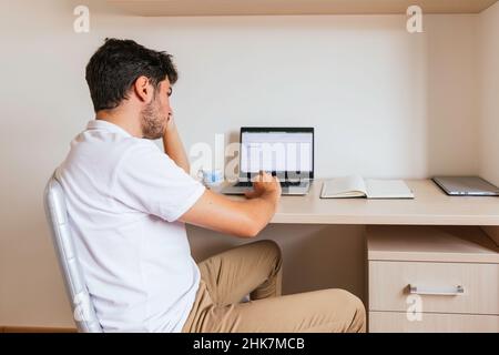 Young man sitting sideways, looking seriously at the computer with one hand on his head. Concept: teleworking or having online classes. Stock Photo