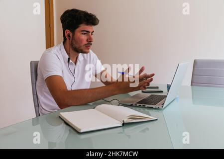 Serious young man explaining with his hands on a work call or an online class, taking notes and talking with his cable headphones. Concept: teleworkin Stock Photo
