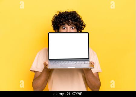 Shocked indian guy, peeking out from behind laptop, looks surprised at camera, stands on isolated orange background, holds an open laptop with blank white screen, copy space, mock-up concept Stock Photo