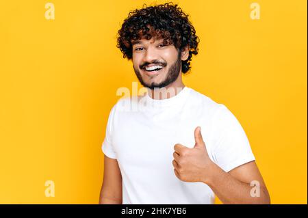 Positive attractive young adult indian or arabian man dressed in basic white t-shirt, showing thumbs up gesture, looks at camera, standing on isolated orange background, smiling happily Stock Photo