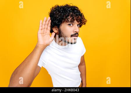 Deafness, gossip concept. Positive Indian man, wearing basic t-shirt with hand near ear concentrated listening rumor, hearing gossip, standing on isolated orange background Stock Photo