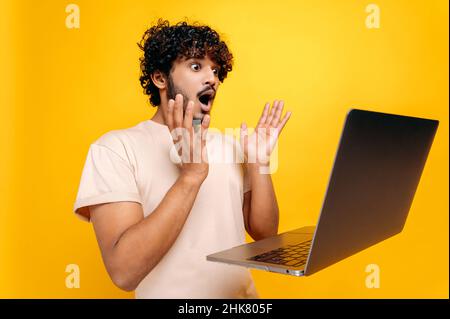 Shocked amazed indian or arabic curly-haired guy, looking in surprise at the laptop screen, saw unexpected news, victory, profit, standing on isolated orange background, gesturing hands.Emotional face Stock Photo