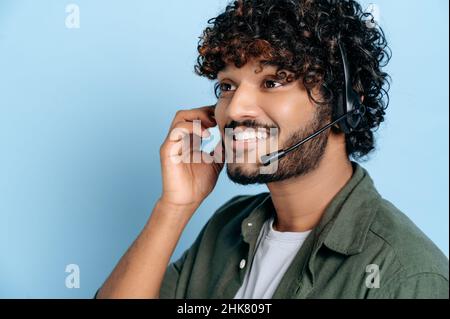 Positive pleasant call center worker, support service operator, expert with headphones, in casual wear, holds distant consultation, standing on blue isolated background, smiles friendly Stock Photo