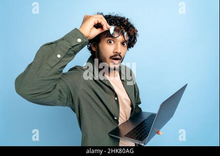 Shocked amazed Indian or Arabian guy, in casual wear, take off glasses, holds an open laptop in hand, looks surprised and confused at the camera, standing on isolated blue background Stock Photo