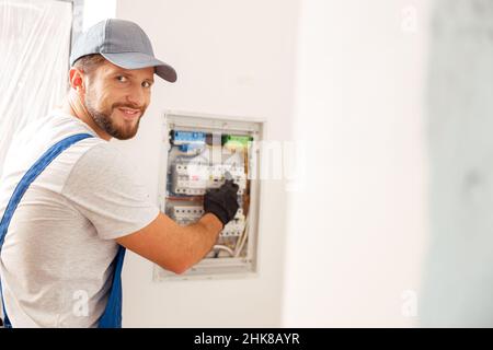 Smiling electrician looking at camera while working with fuses at the circuit breaker control cabinet on the wall Stock Photo