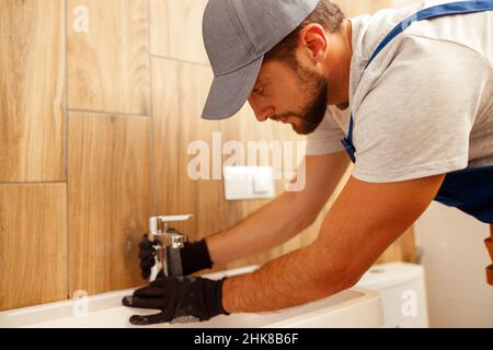 Serious plumber, male worker in uniform installing tap or bathroom faucet Stock Photo