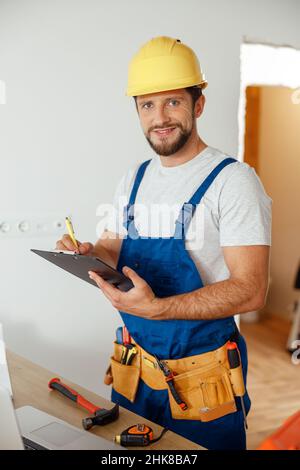 Joyful handyman worker, builder in uniform and hard hat looking at camera, making notes on a clipboard while standing indoors, getting ready for Stock Photo