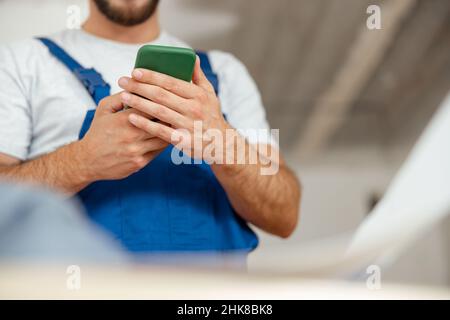 Closeup of hands of repairman in workwear texting, using smartphone, standing indoors during renovation work Stock Photo