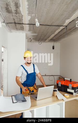 Happy repairman worker in uniform wearing tool belt and hardhat holding hammer and smiling at camera while standing indoors Stock Photo