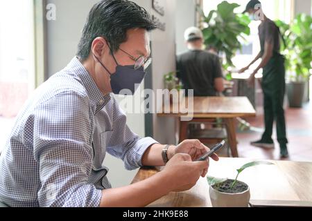 Asian man with face mask on using his phone, text messaging in a restaurant. Stock Photo