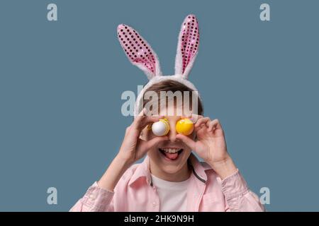 Easter boy funny portrait. Happy boy with Easter eggs Caucasian child holding colorful Easter eggs in front in his hands and smiling with different facial expressions, pink shirt on blue background Stock Photo