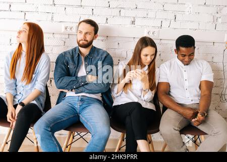 Tired young diverse multiethnic job candidates in casual clothes waiting interview with hr, sitting in queue line row on chairs. Stock Photo